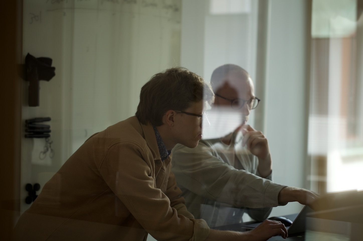 Två personer samarbetar vid en laptop i mötesrum, fotograferade genom glasvägg.