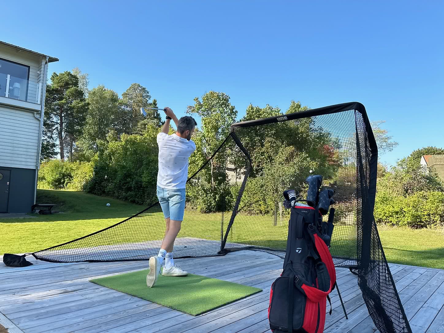 En person svingar en golfklubba på en hemmabana med ett träningsnät i bakgrunden. En golfbag står bredvid.