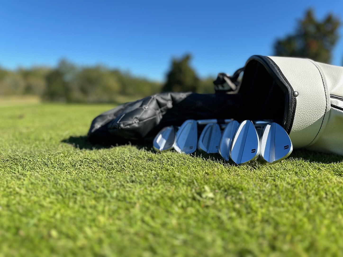 En närbild av glänsande golfklubbor som ligger utspridda framför en golfbag på en grön fairway under klarblå himmel.