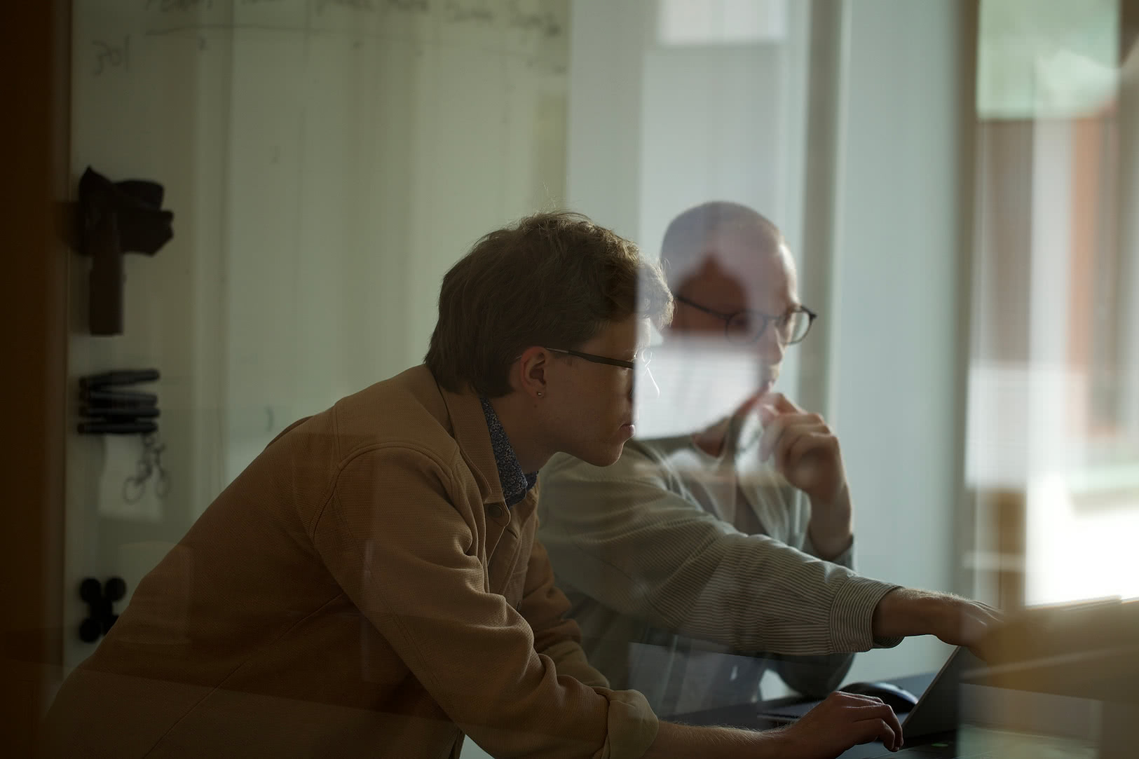 Två personer samarbetar vid en laptop i mötesrum, fotograferade genom glasvägg.