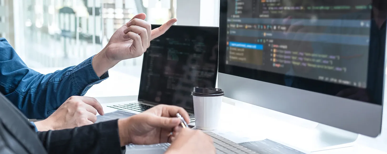 Close-up of a workspace with a laptop and a screen, two people sit in front of the computer interacting with each other.