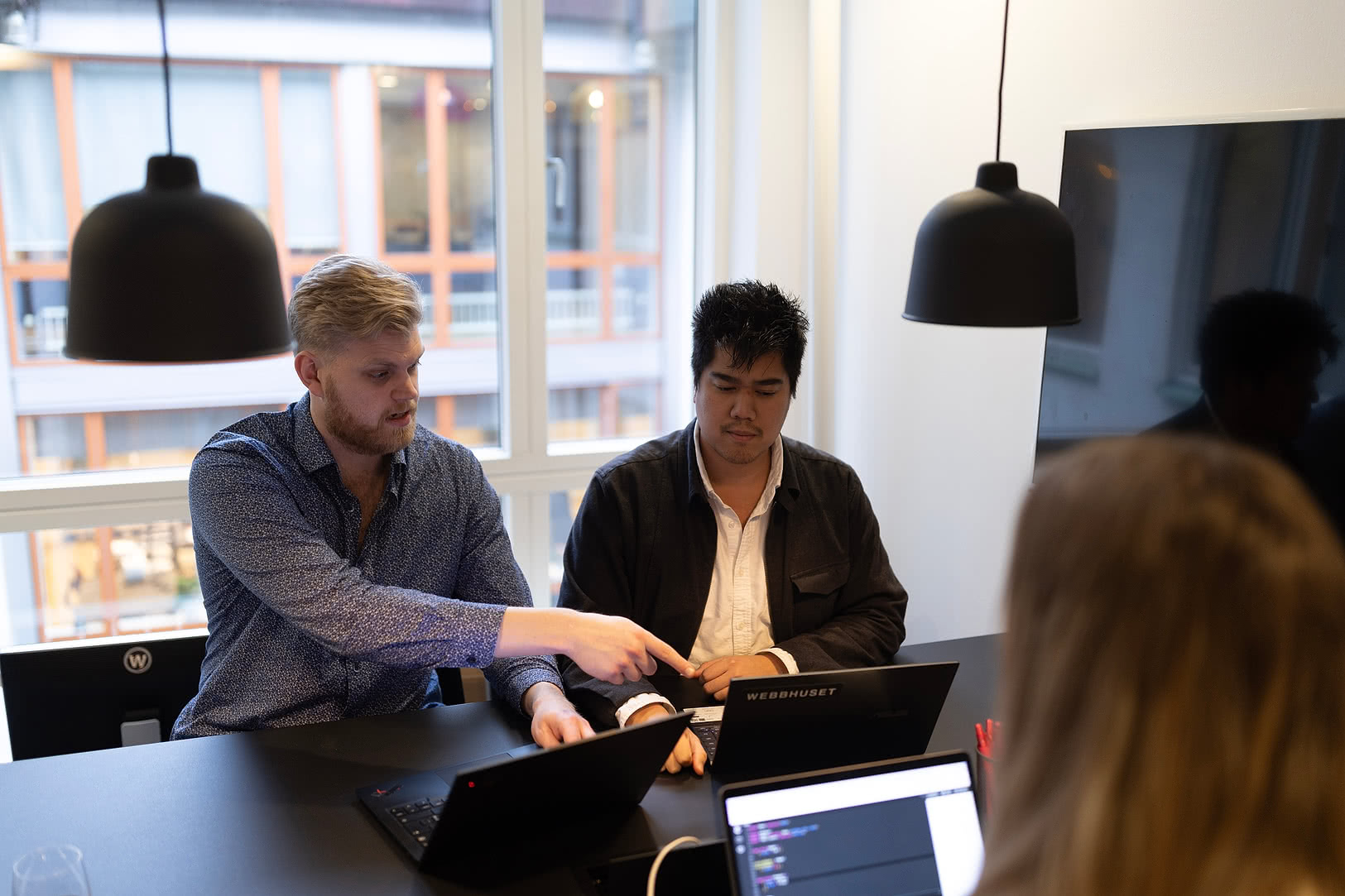 Två medarbetare sitter framför en laptop och arbetar tillsammans. En tredje person sitter mittemot med ryggen vänd från kameran och kollar på medarbetarna. 
