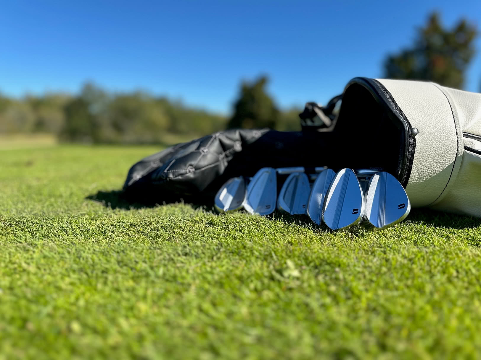 En närbild av glänsande golfklubbor som ligger utspridda framför en golfbag på en grön fairway under klarblå himmel.