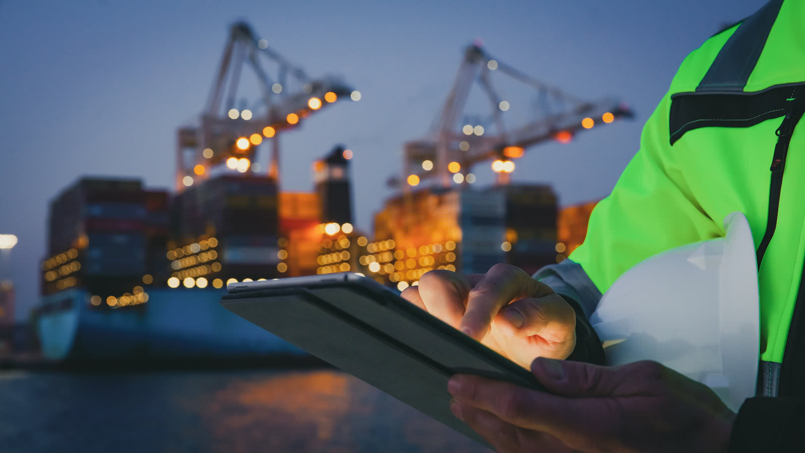 Person in a high-visibility jacket using a tablet in a port at night, with container ships and cranes in the background.