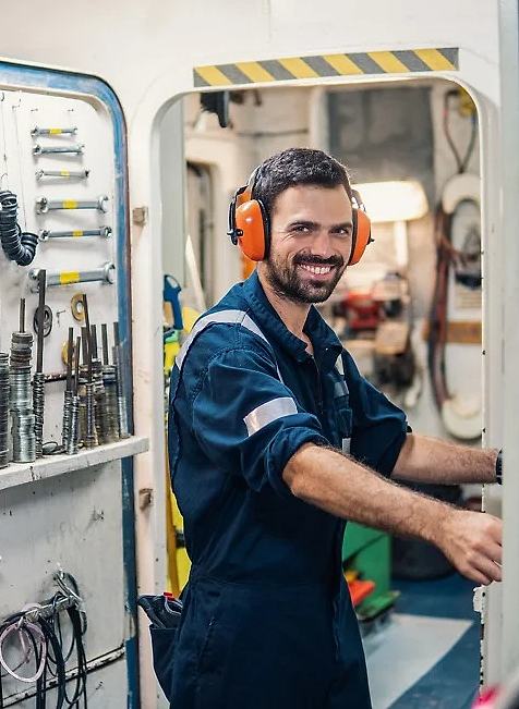 Smiling sailor in a blue overall and ear protection stands in an engine room aboard a ship, surrounded by tools and equipment.