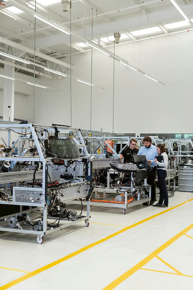 Three workers in the manufacturing industry review production data on a laptop at an assembly station with vehicle components in a modern factory facility.